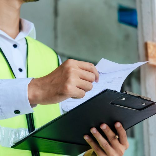 engineer or home inspector in green reflective jacket checking review document and inspecting with clipboard at construction site building interior, construction, contractor and engineering concept